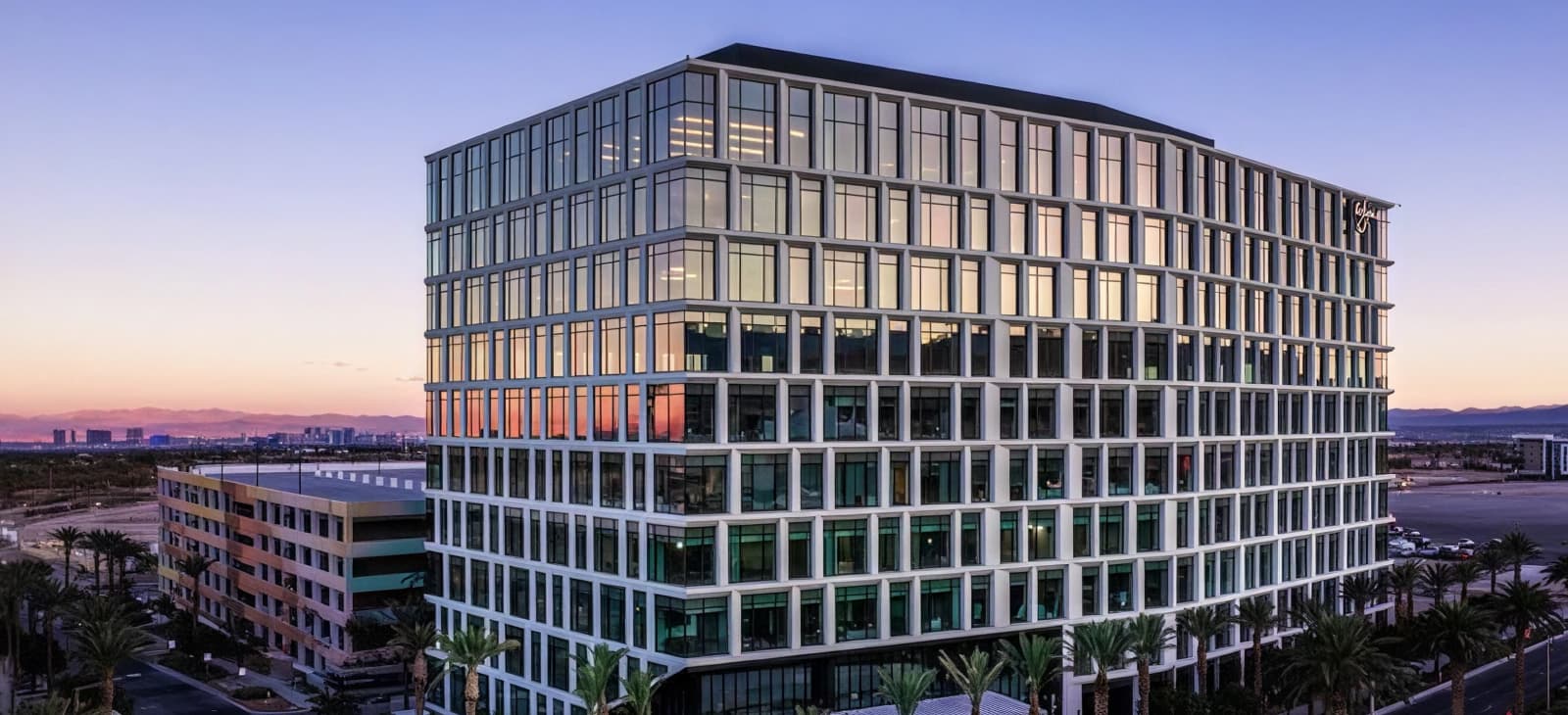 Modern Glass Office Building At Dusk With Illuminated Interior And City Skyline